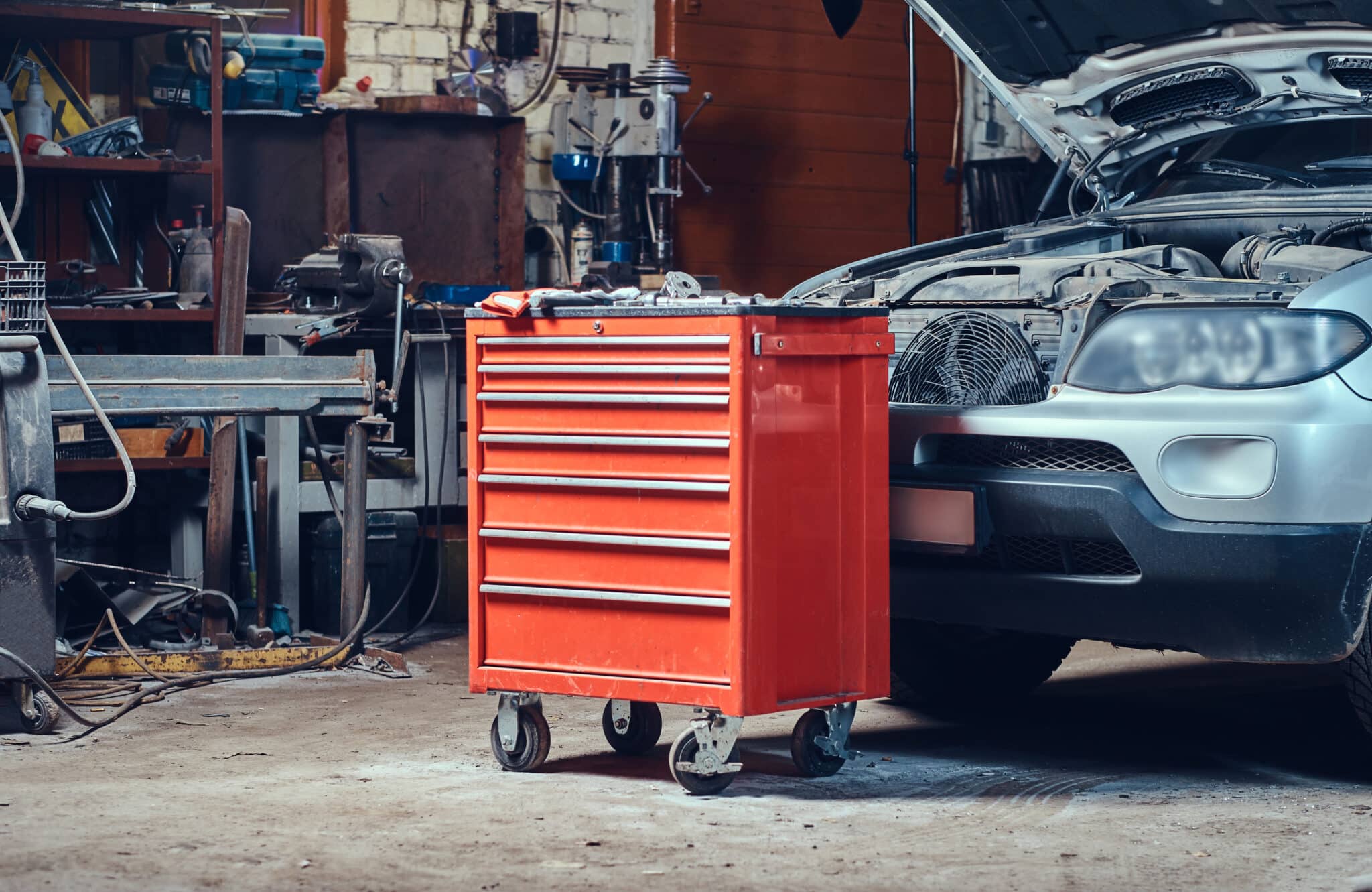 Servante d'atelier rouge à tiroirs sur roulettes, devant une voiture au capot ouvert dans un garage