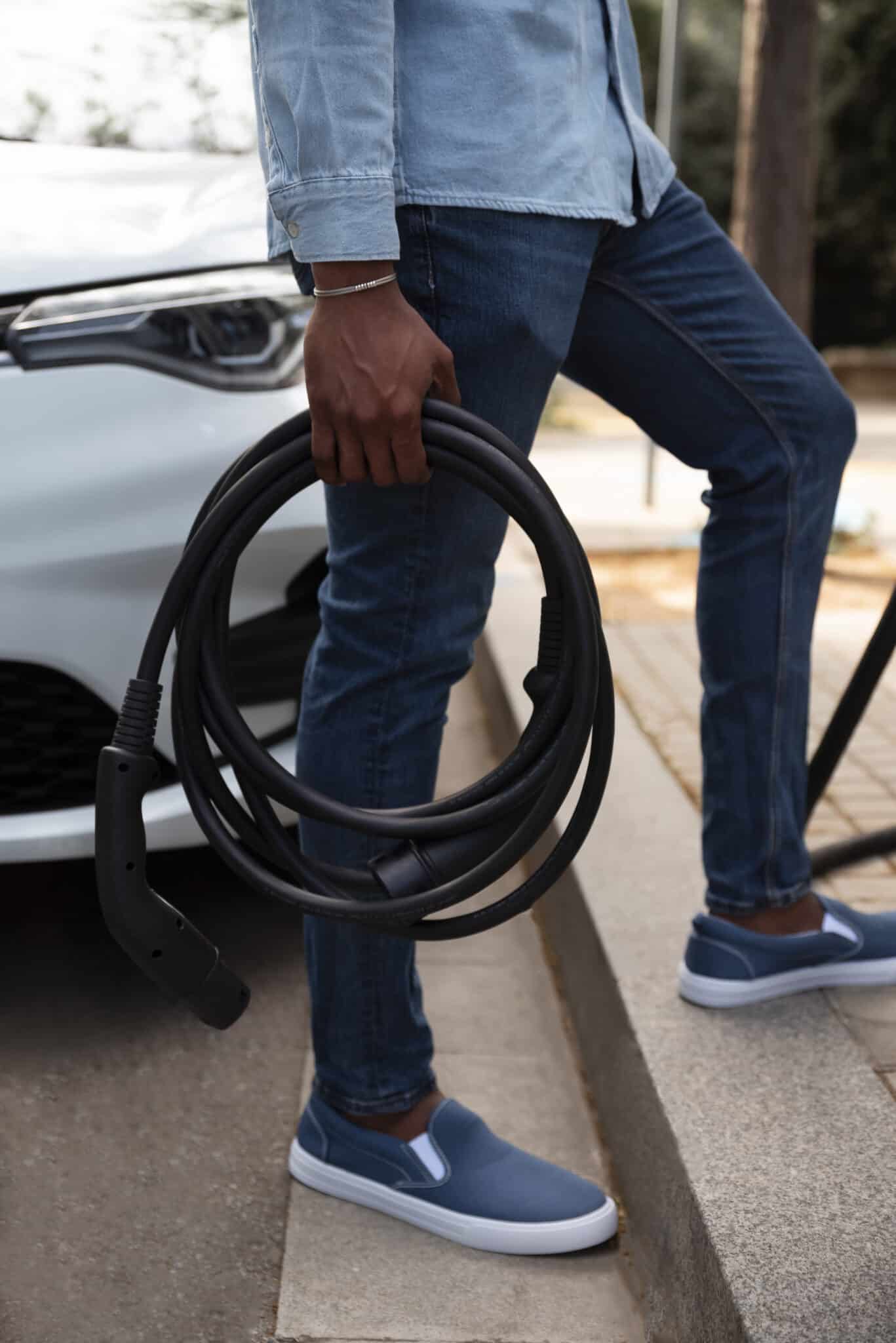 Homme en jean tenant un câble de recharge noir enroulé près de l'avant d'une voiture blanche