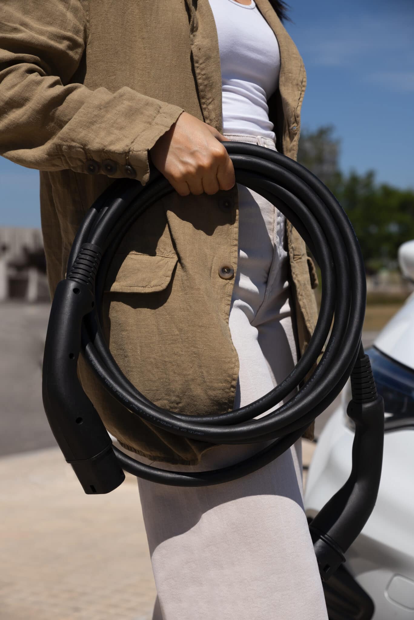 Personne en veste beige tenant un câble de recharge noir enroulé, près d'une voiture blanche